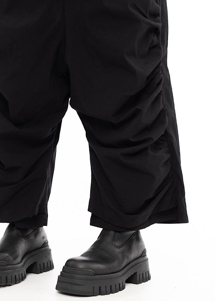An up close of A Model wearing a drop-crotch Trouser in Black. With a kick-flare leg and ruched side seams. Showing the details around the ankle.