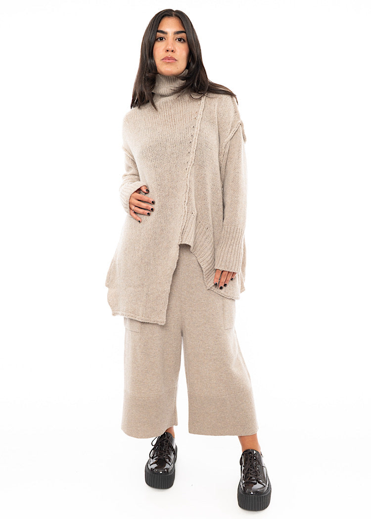 A full frame photo of A Model wearing a light beige knitted Trouser. Paired with a matching Oversized pullover and Brown patent Shoes.