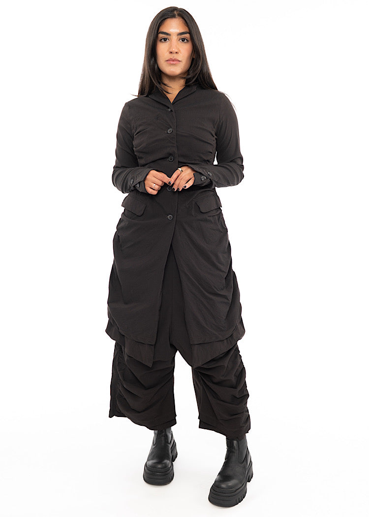 A full frame photo of A Model wearing a minimal design techno coat and matching trousers, in a dark off-Brown tone. Paired with a long Boot.