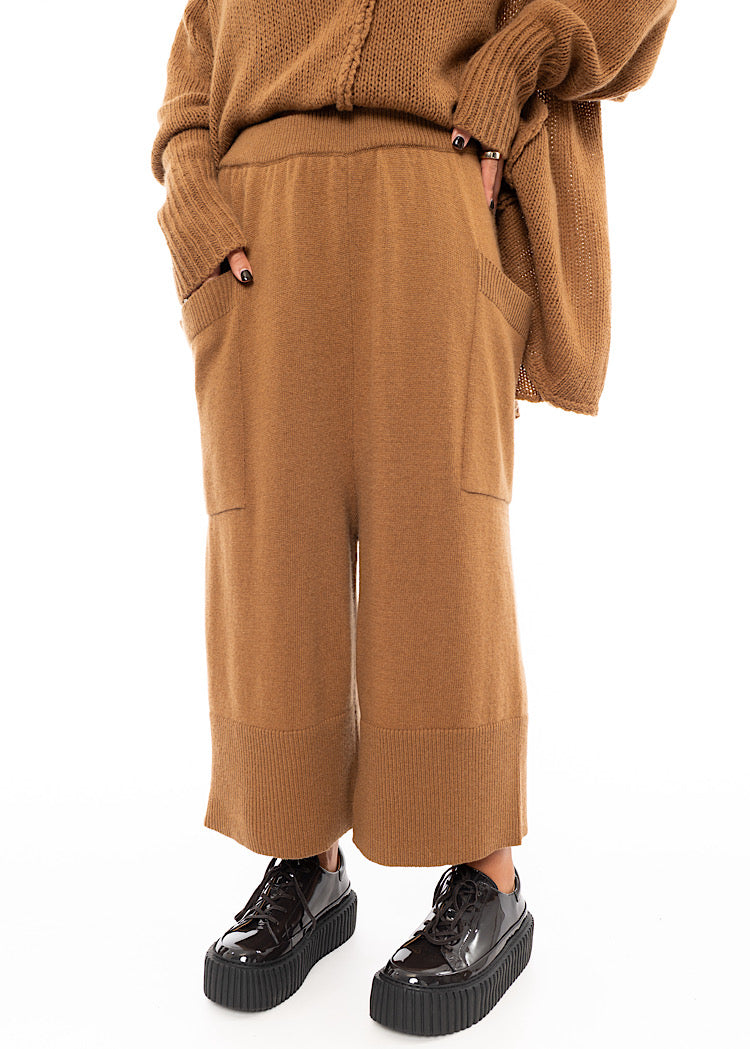 A Model wearing a Knitted pair of off-orange Trousers, paired with a matching pullover and patent shoes.