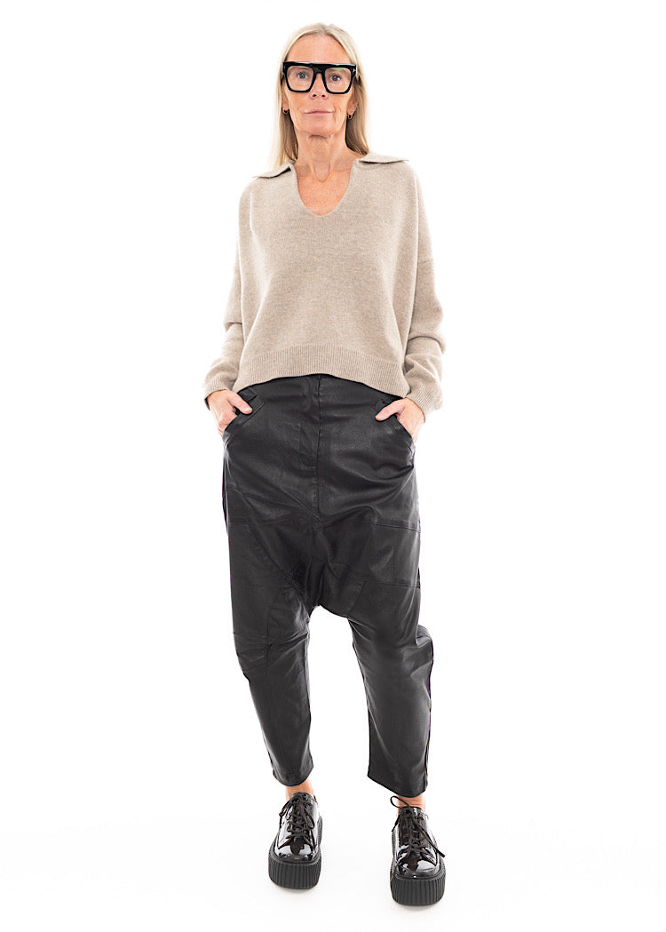 A full frame photo of A Model wearing a Structured Drop-Crotch Black Leather Trouser, disclosing the pockets. Styled With a patent Brown Shoe and a creme