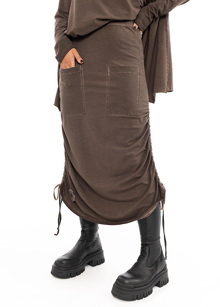 A Model wearing a Chocolate Brown skirt with distinctive details. Focusing on two square pockets and drawstring effects. Styled with matching Brown pullover and Black boot.