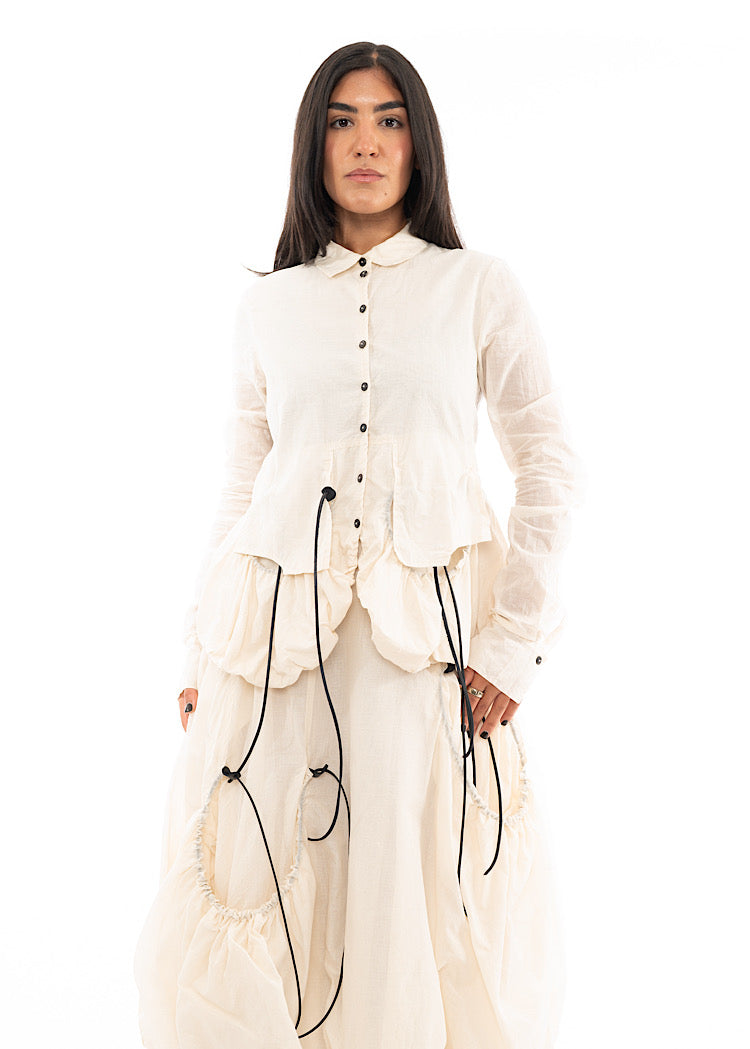 A Model wearing a sculptural Jacket with a Skirt in the matching light creme colour. Focusing on the layered Black drawstrings and fitted buttons.