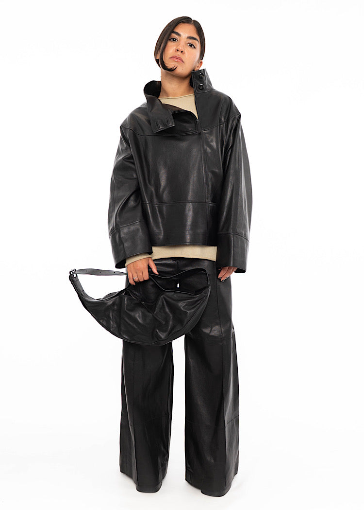 A Model wearing an oversized Black Leather pullover with an open unique collar. Styled with matching leather trousers, a beige pullover underneath and a slouchy matching leather bag.