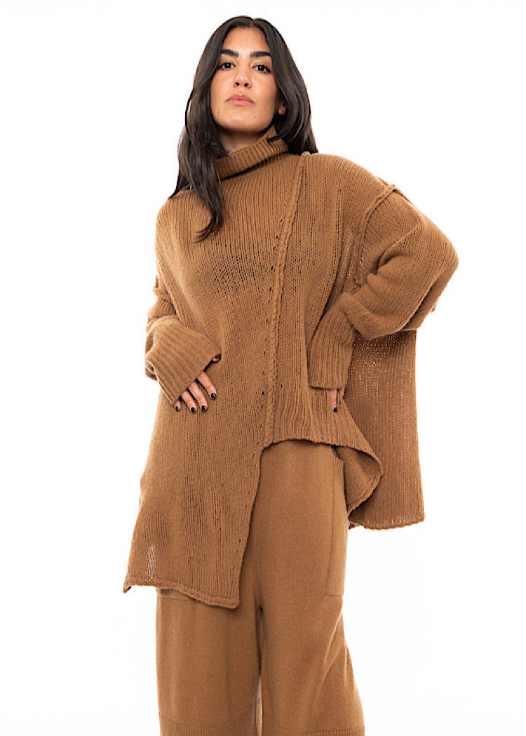 A Model wearing a light brown knitted Oversized Pullover with distressed pieces dangling down. Paired with a matching coloured Trouser.