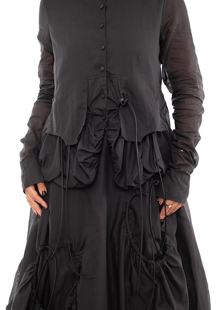 An up close off A Model standing to a side wearing a sculptural Jacket with a matching Skirt in the same washed Grey colour.