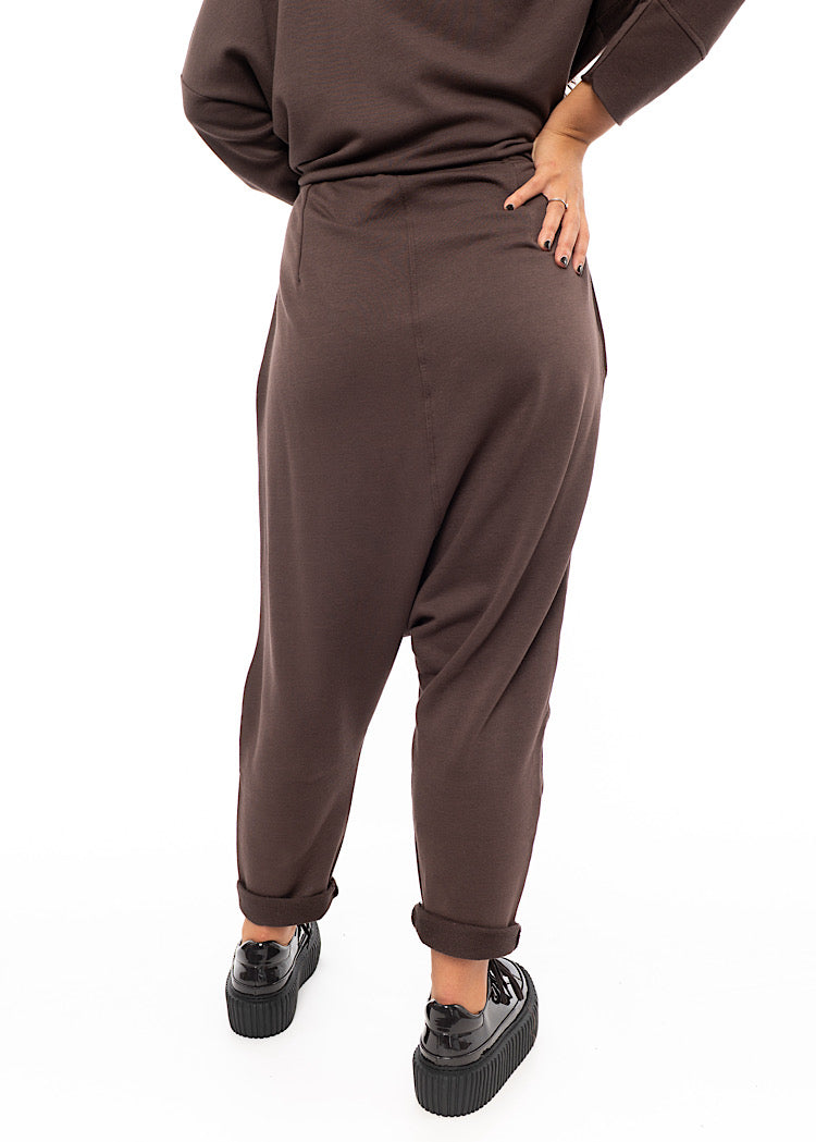 A Model standing facing the back wearing a plum toned drop-crotch trouser with a slight rolled up effect on the Ankle.