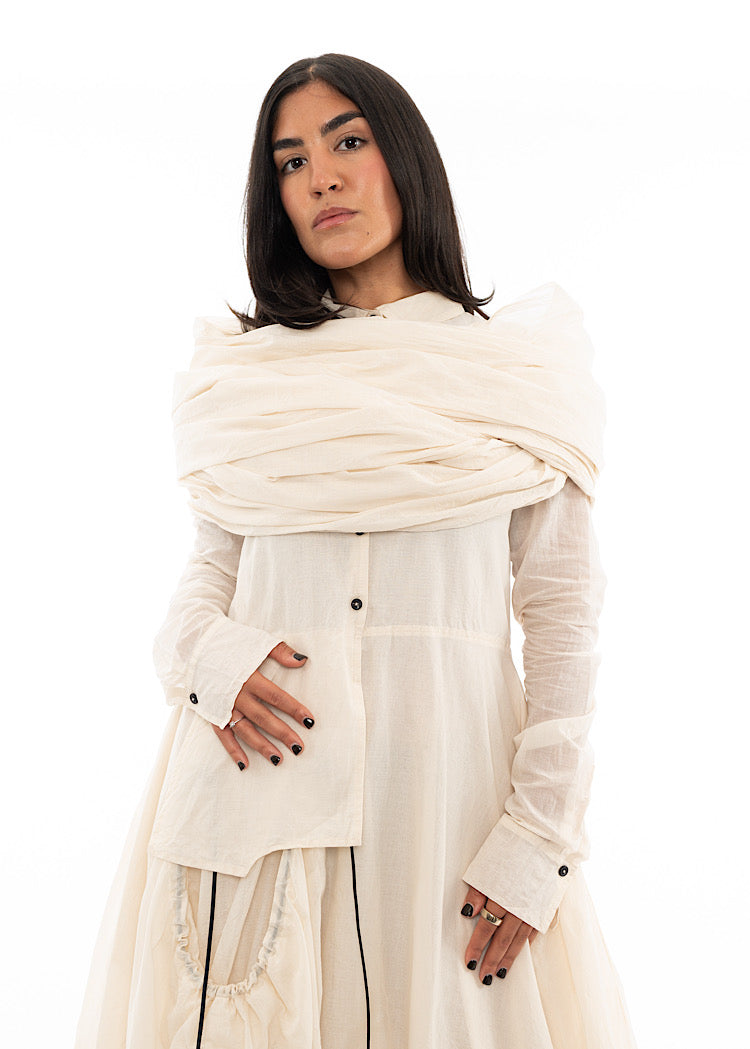 A Model wearing an oversized Scarf wrapped around her shoulders adding volume to the matching Dress in a light creme colour.