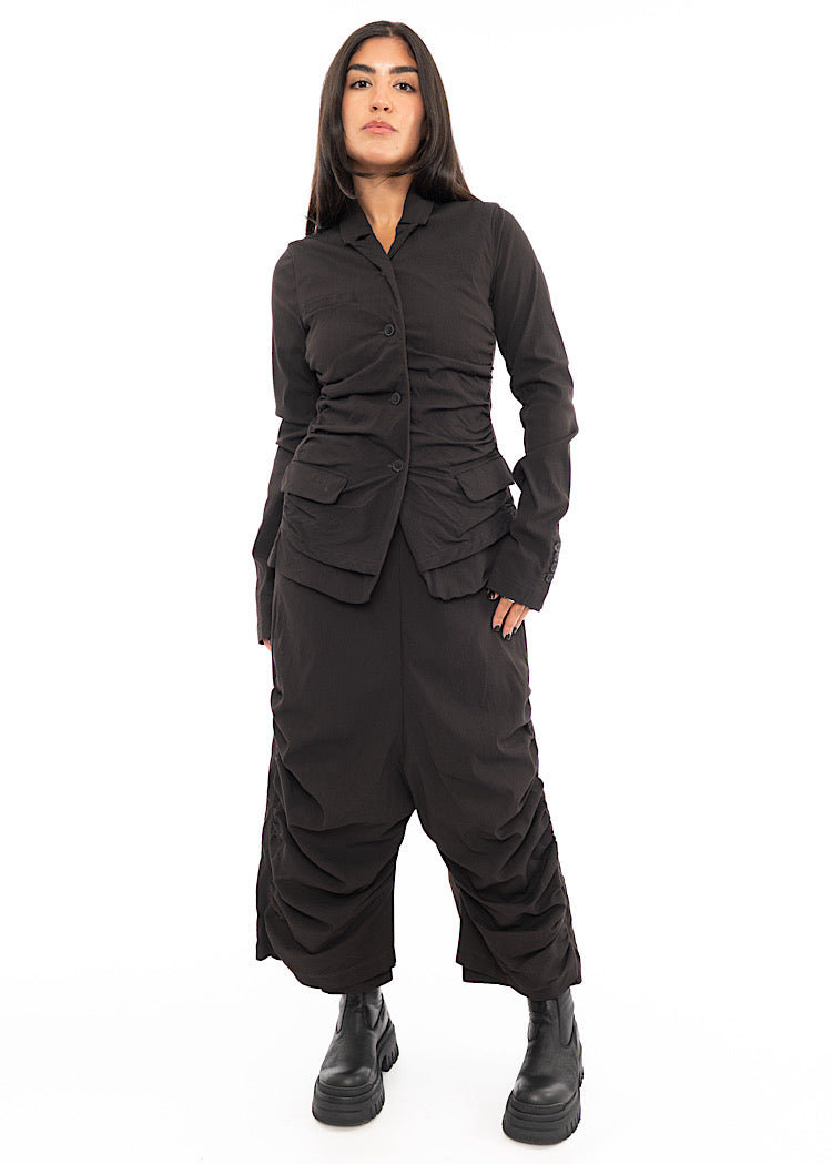 A full frame photo off A Model wearing a detailed techno Jacket and matching trousers, in a dark off-Brown colour. Paired with a Black chunky boot.