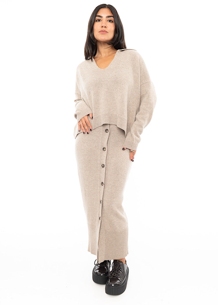 A full frame photo of A Model wearing a light beige completely buttoned up Skirt. Paired with a matching pullover and patent Brown Shoe.