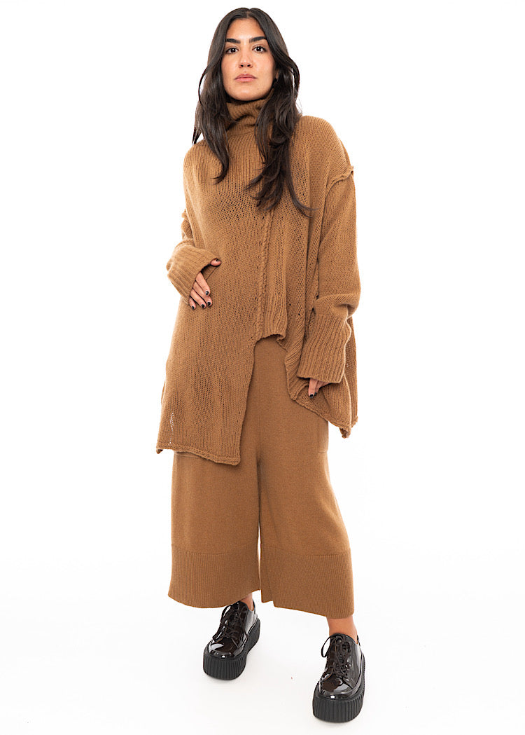 A Model wearing an off-orange knitted Oversized Pullover with distressed pieces dangling down. Paired with a matching coloured Trouser.