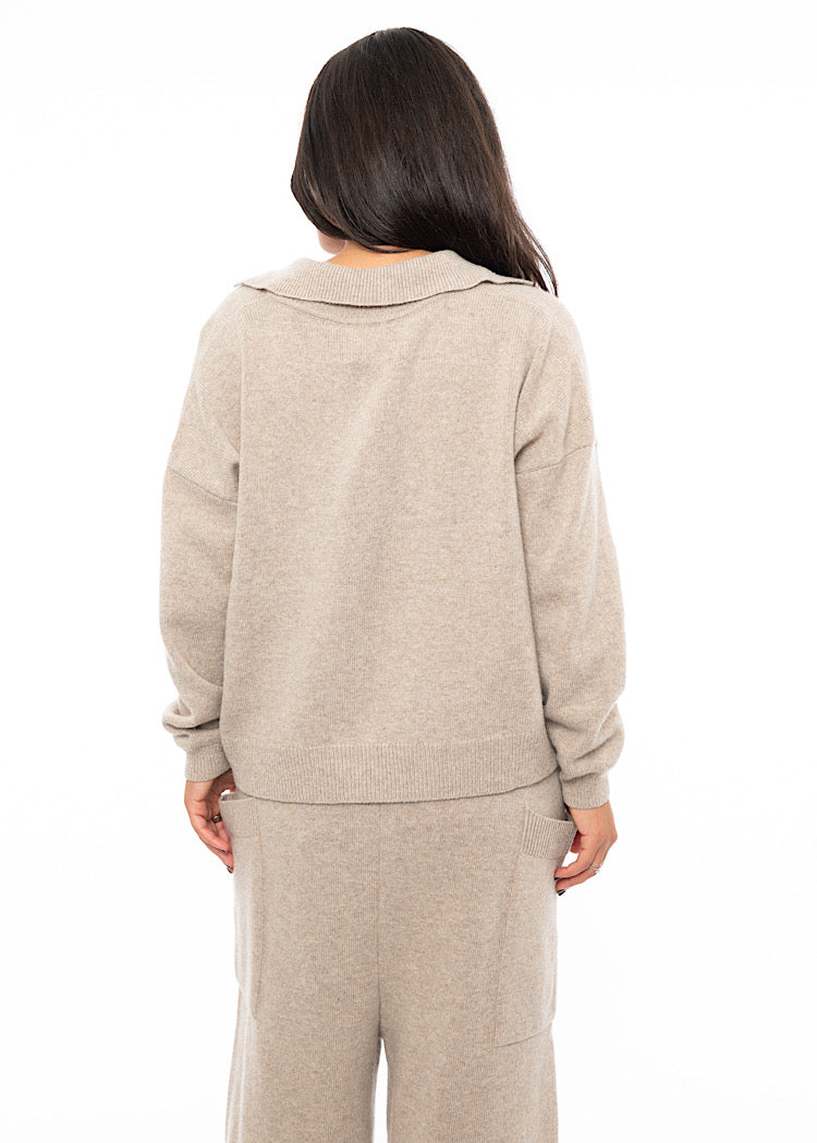 A Model standing facing the Back wearing a light Beige knitted pullover with a ribbed hem, a deep V-neckline and a relaxed collar. Showing the pieces length.