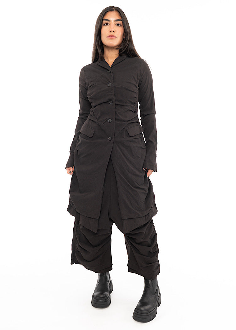 A full frame photo of A Model wearing a detailed techno coat and matching trousers, in a dark off-Brown tone. Paired with a chunky Boot.