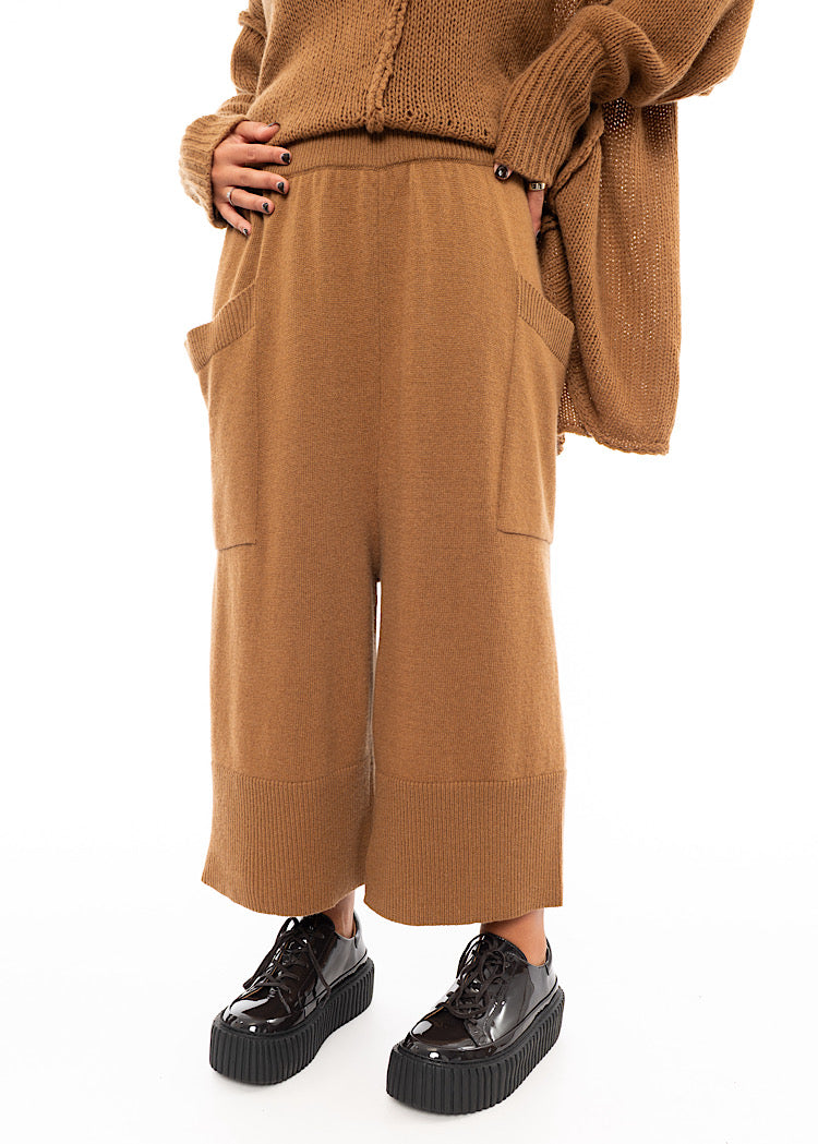 A Model wearing a Knitted pair of off-orange Trousers, paired with a matching pullover and patent shoes.Showing the ribbed hem.