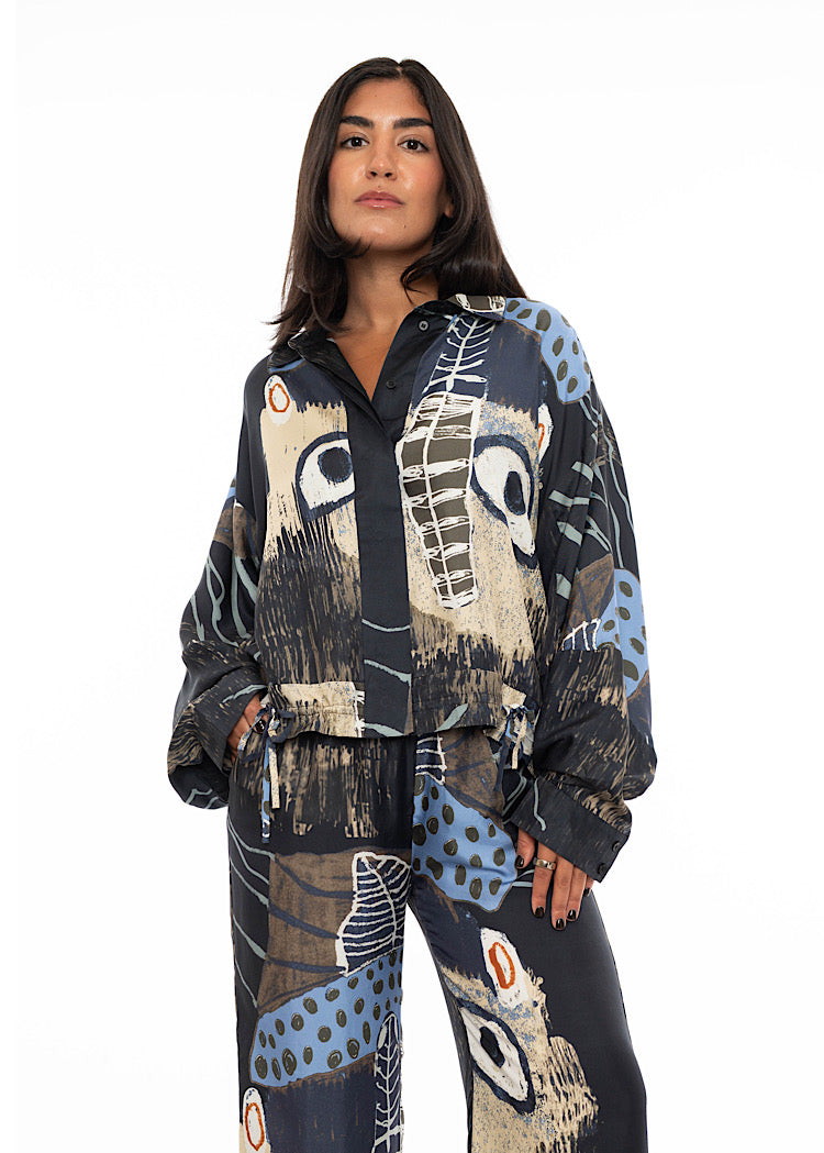 A model wearing an oversized silk shirt with multicoloured print throughout. With hidden buttons to fasten and styled with matching silk trouser.