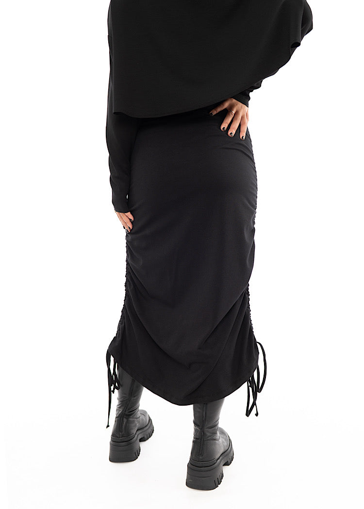 A Model standing facing the back wearing a Black drawstring Skirt, styled with a cropped Black Pullover and Chunky Black Boot.