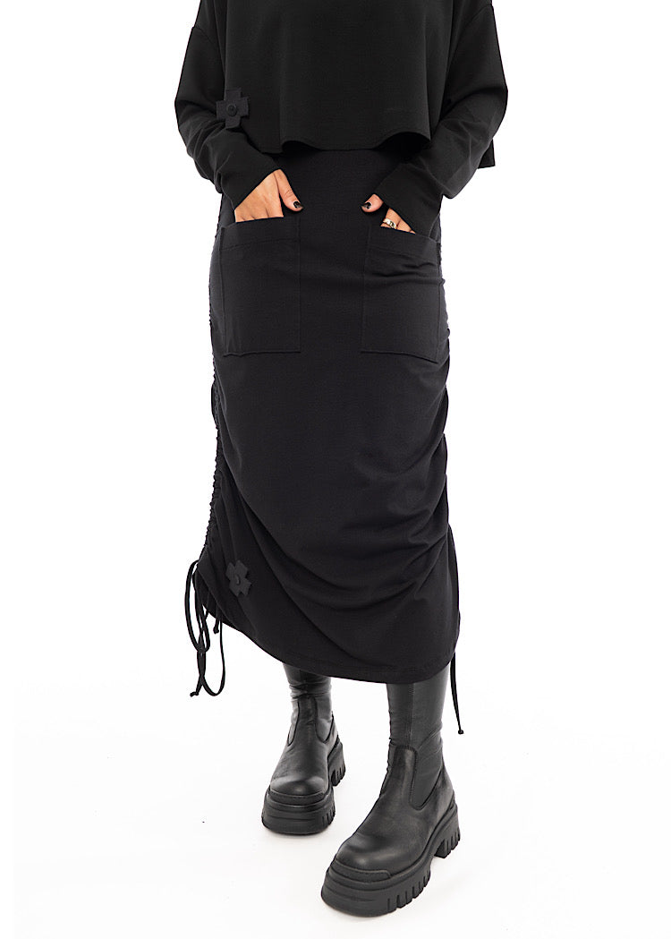 A Model wearing a Black drawstring Skirt, styled with a cropped Black Pullover and Chunky Black Boot.Disclosing the hidden pockets.