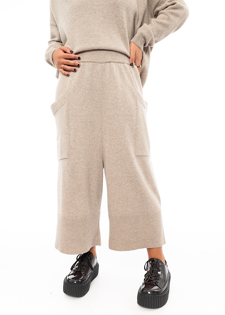 A Model wearing a light beige knitted Trouser. Paired with a matching pullover and Brown patent Shoes.