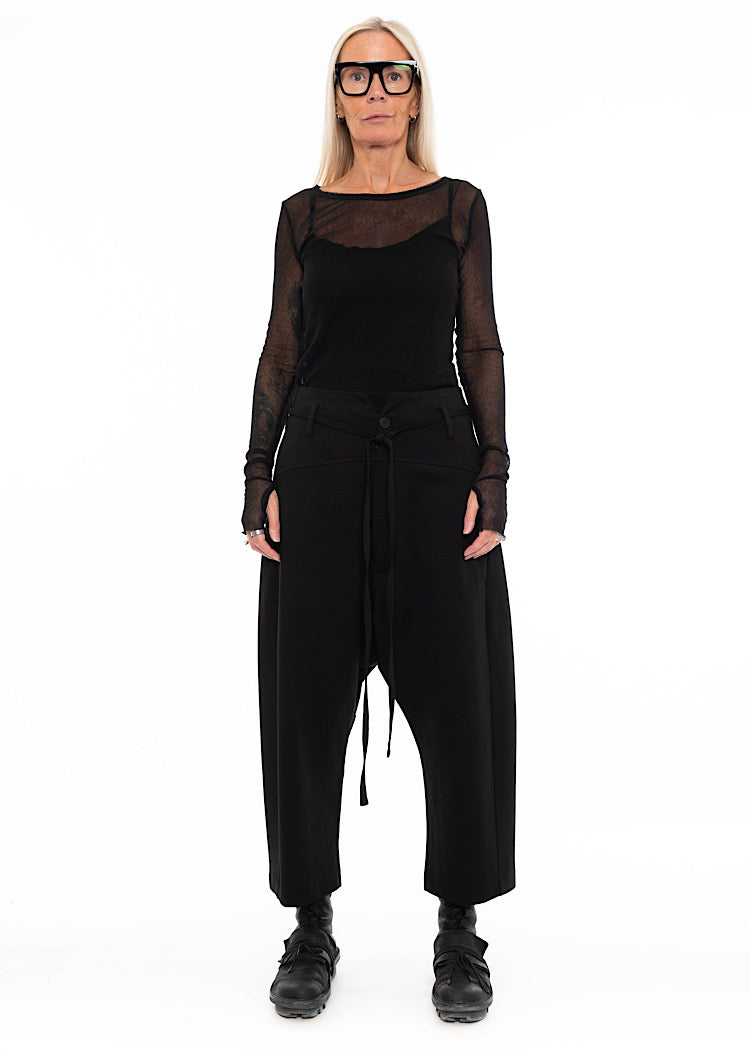 Model wearing black drop-crotch trousers with wide leg and drawstring detail, styled with mesh-sleeve top with combat boots.