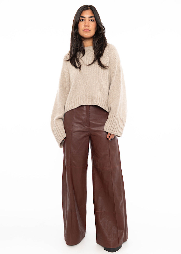 Model wearing Burgundy wide leg leather trousers with a zipper and button to fasten. Styled with an oversized warm beige pullover with ribbed detailing and black high heels.