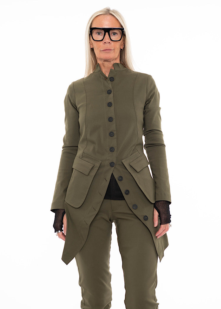 Model wearing a tailored khaki green longline blazer with asymmetric hem and black button fastening, styled with matching trousers and mesh sleeves.