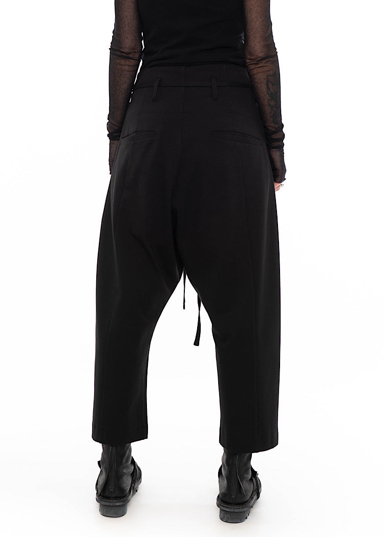 Rear view of Model wearing black drop-crotch trousers with wide leg and drawstring detail, styled with mesh-sleeve top and combat boots.