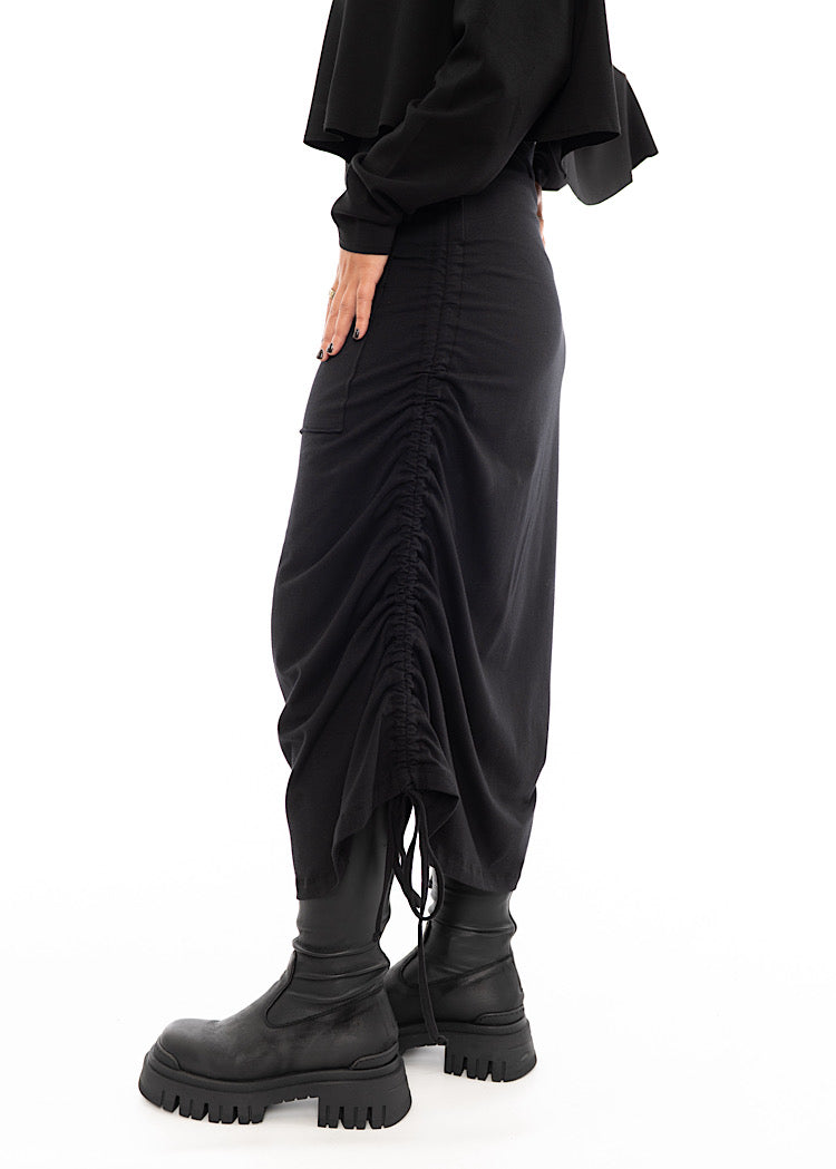A Model standing to the side wearing a Black drawstring Skirt, styled with a cropped Black Pullover and Chunky Black Boot. Showing the length of skirt.