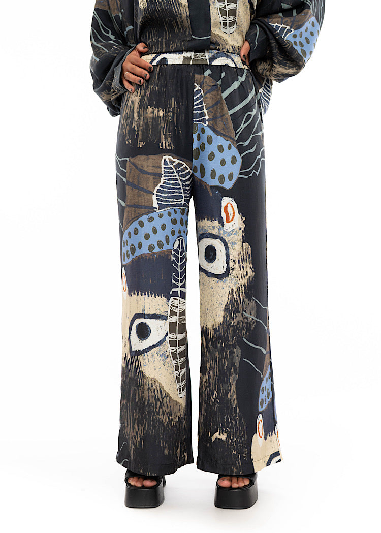 A close up of a model wearing wide leg Silk trousers with a multicoloured pattern throughout. Showing the high waisted length and flare style trouser.