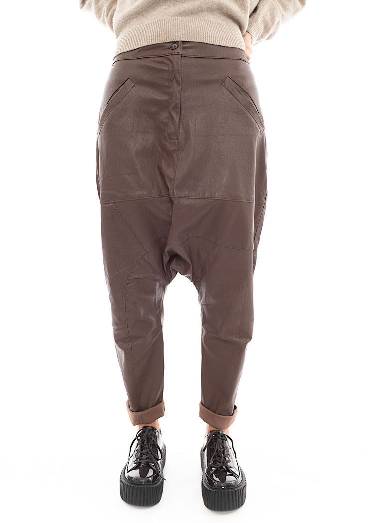 A Model wearing a Structured Drop-Crotch Brown Leather Trouser.Styled With a patent Brown Shoe and a creme pullover.