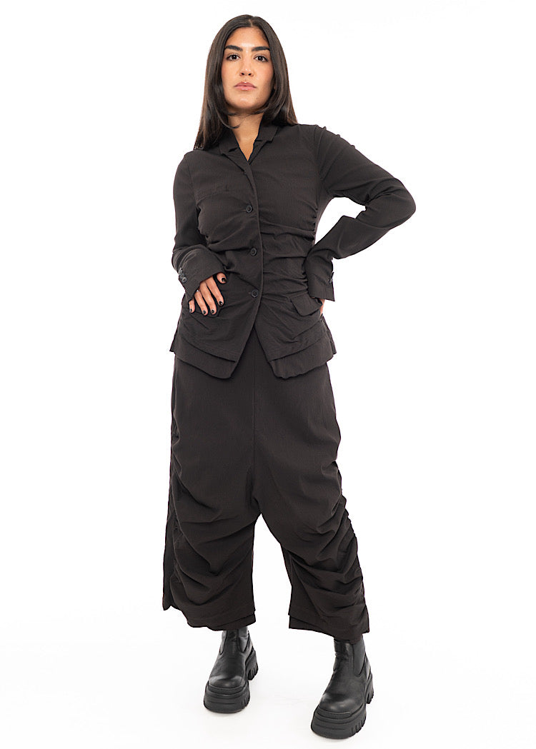 A full frame photo off A Model wearing a detailed techno Jacket and matching trousers, in a dark off-Brown colour. Paired with a Black chunky boot.