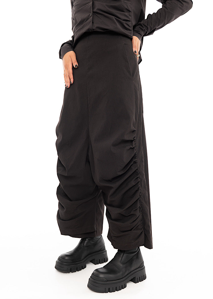 A Model standing slightly to an Angle, wearing a drop-crotch Trouser in a washed dark Brown colour. Showing the straight fit of the waist.