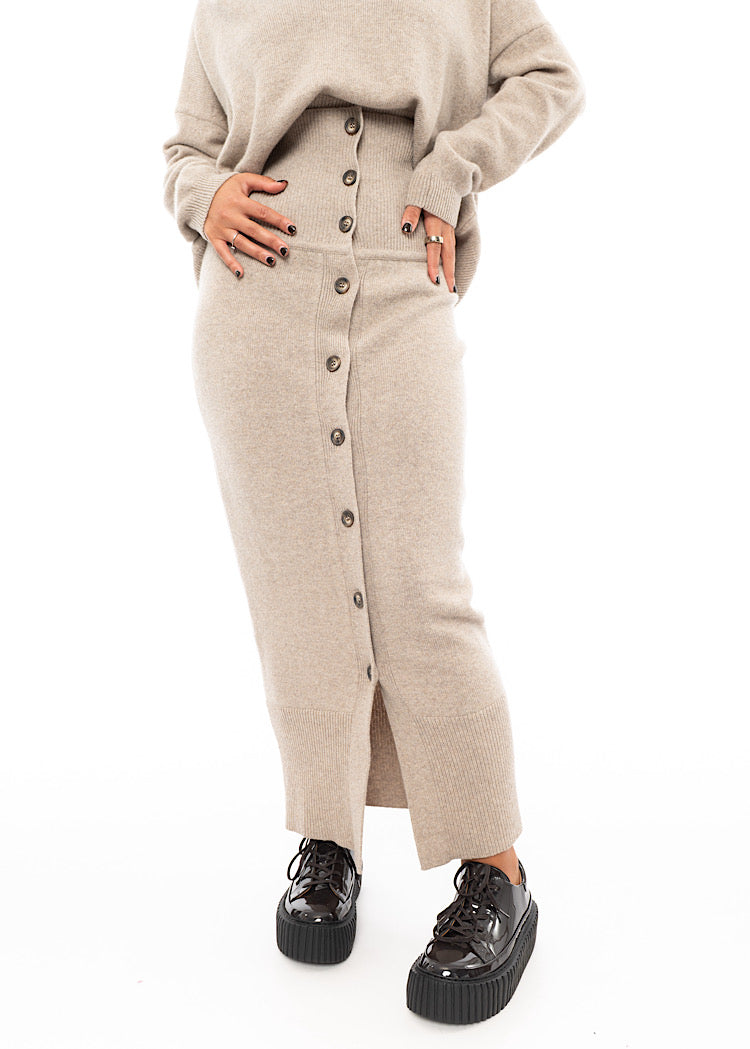 A Model wearing a light beige completely buttoned up Skirt, paired with a matching Pullover and a patent Brown Shoe