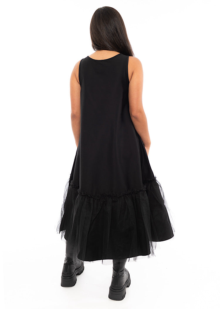 A Model standing facing the back wearing a fitted Black dress which bottom hem turns into a floaty mesh skirt. Showing the dress from behind.