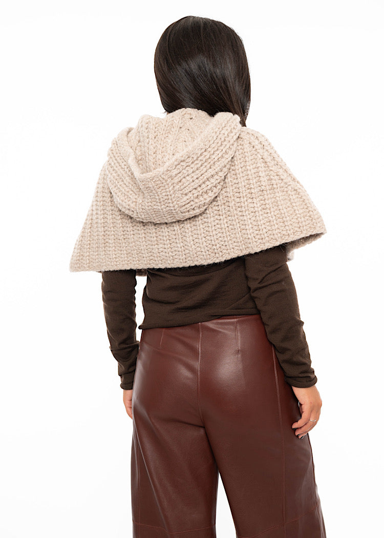 A model standing facing the back wearing an oversized knitted shawl with a hood in creme with beige minimal buttons to fasten. Showing the how the hood sits on the back of piece.