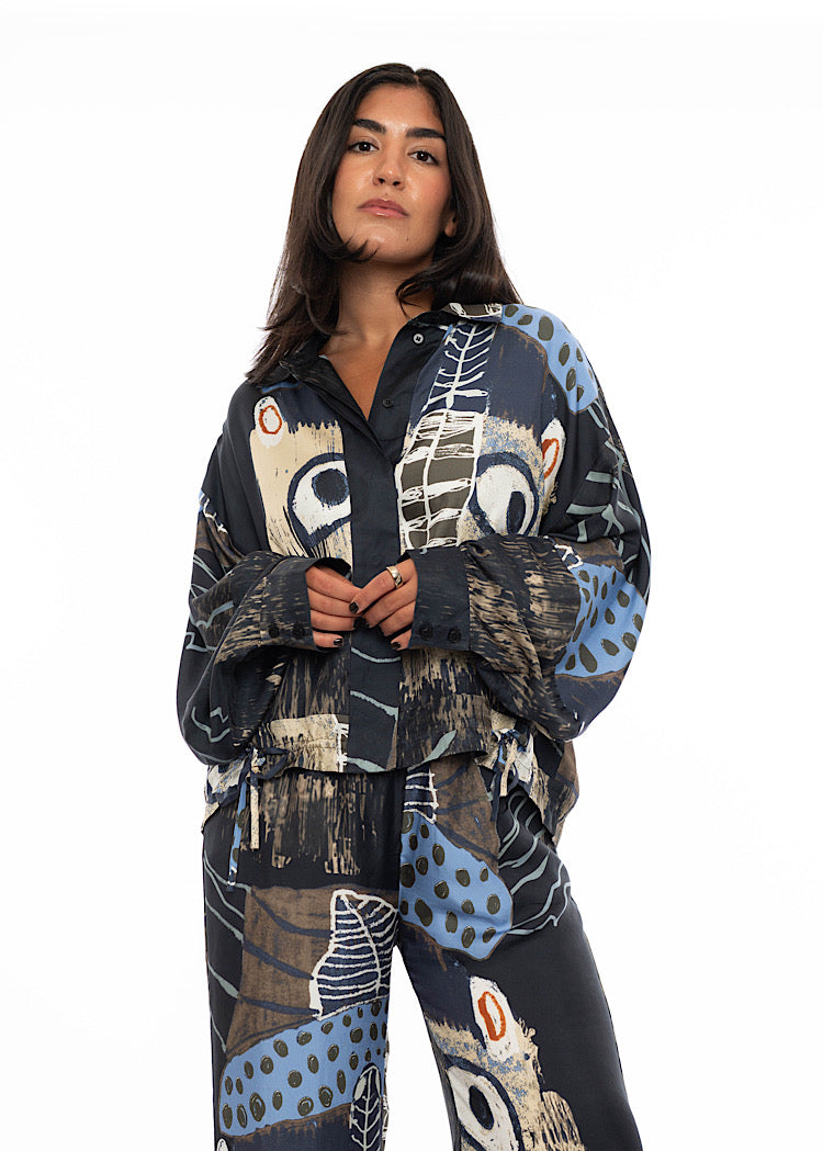 A model wearing an oversized silk shirt with multicoloured print throughout. Showing the floaty cuff detailing on the arms .Paired with a matching silk wide leg trouser to add volume paired with black high heels.