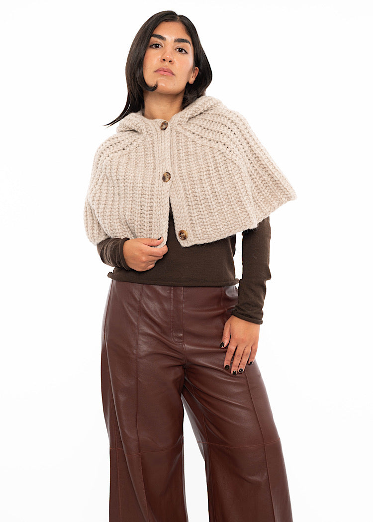 A model wearing an oversized knitted shawl with a hood in burgundy. Styled with a fitted black pullover and Burgundy leather jeans. Showing the minimalist buttons and the length of shoulders.