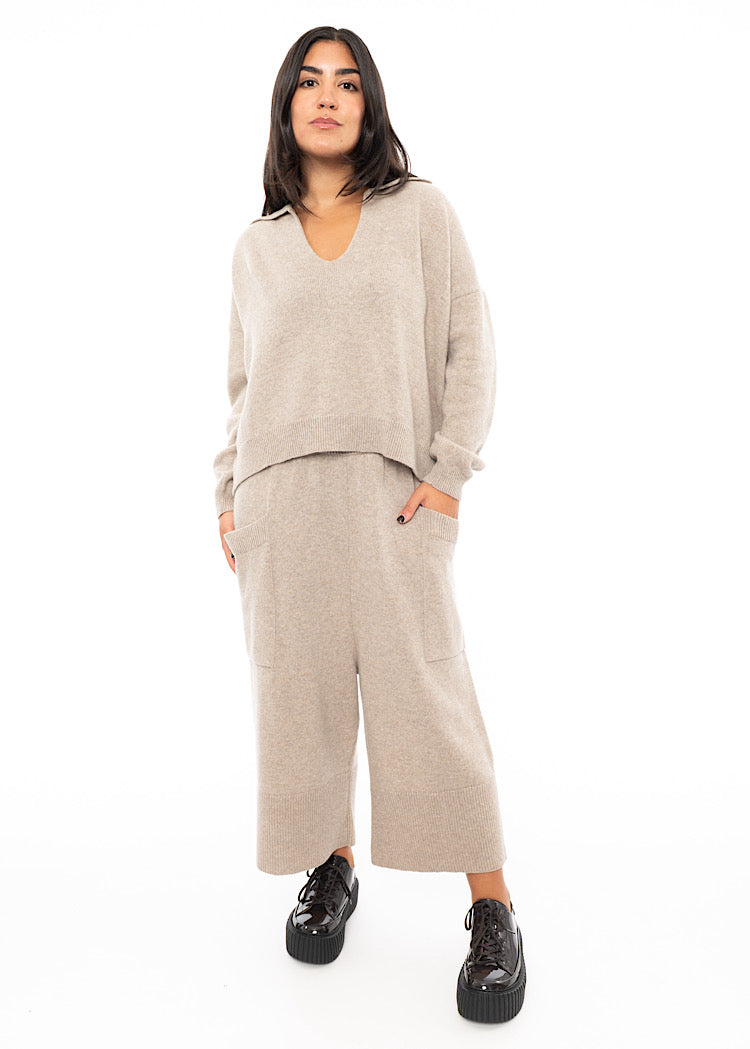 A Full frame photo of A Model wearing a light Beige knitted pullover with a deep V-neckline.Styled with a matching Trouser and patent Brown Shoes.