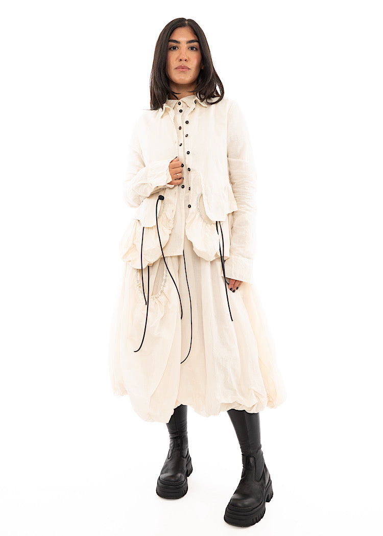 A full frame photo of A Model wearing a bubble skirted Dress in a creme colour. Focusing on the flap pocket, paired with a Black Boot and matching Jacket.