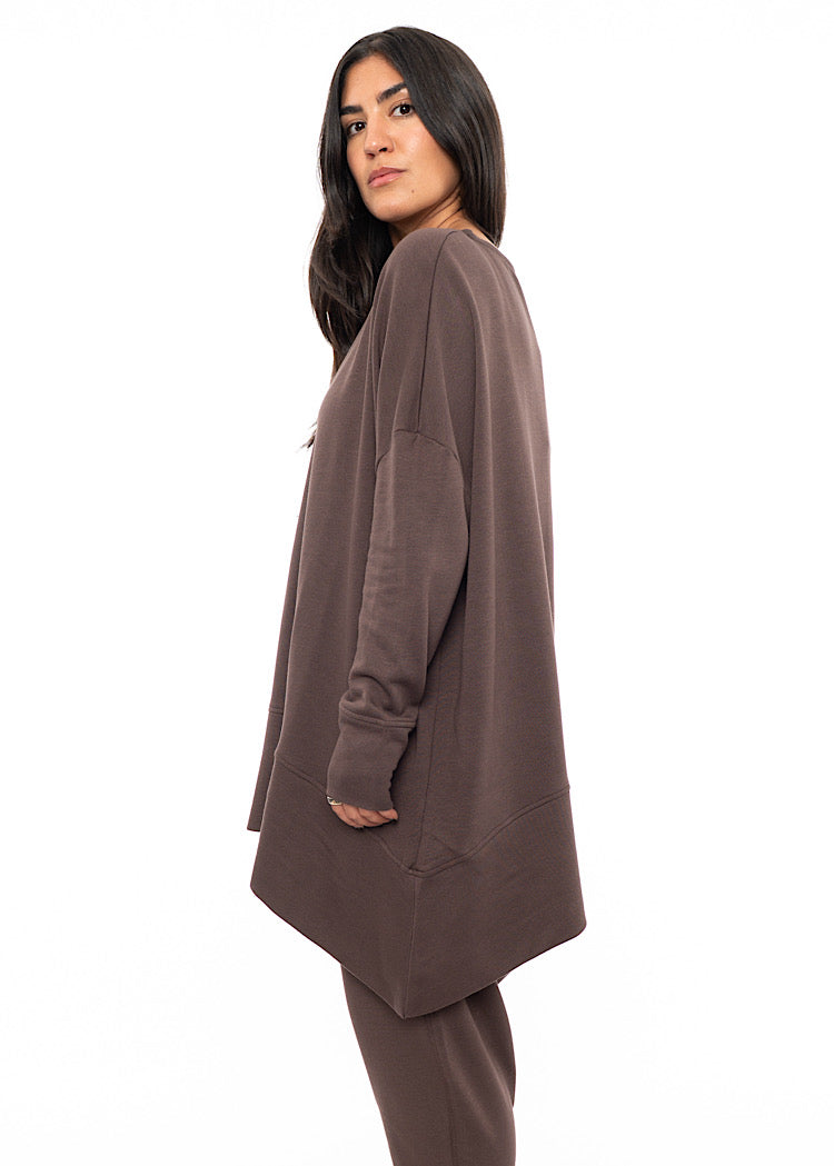 A Model standing to a side wearing an oversized Plum toned Pullover, paired with a Matching Trouser. Showing the pieces long length of arm and body.