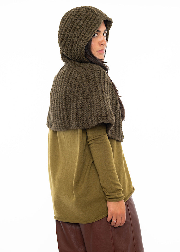 A Model facing towards the back wearing an oversized knitted shawl in dark green. Showing how the hood looks up.