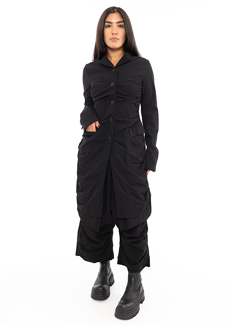 A full frame photo of A Model wearing a detailed techno coat and matching trousers, in a Black colour. Showing the disclosed pockets.