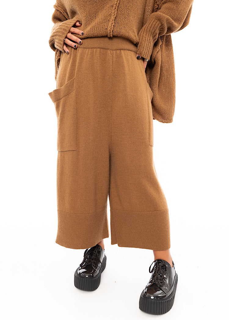 A Model wearing a Knitted pair of off-orange Trousers, paired with a matching pullover and patent shoes.