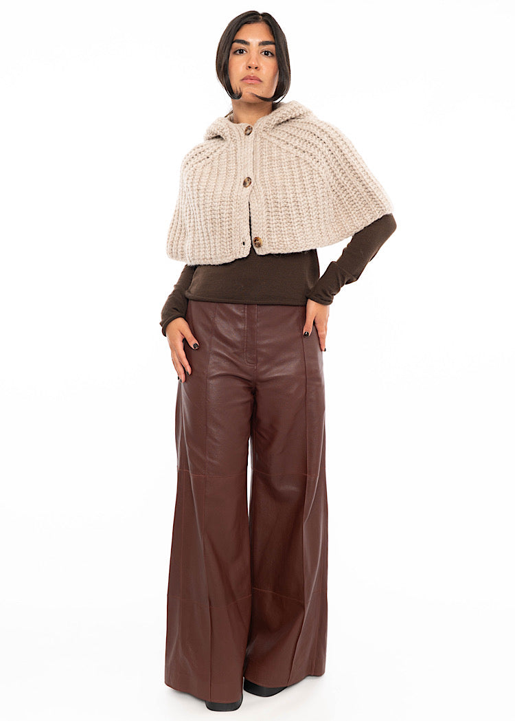 A model standing wearing an oversized knitted shawl with a hood in creme with beige minimal buttons to fasten. Styled with Burgundy leather trousers and a brown fitted pullover.