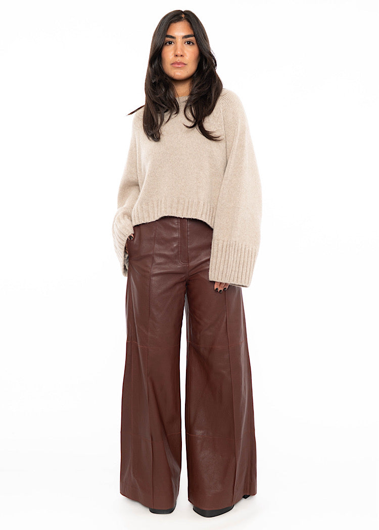 Model wearing an oversized beige pullover with ribbing at the hemline and cuffs. Styled with wide leg leather trousers in burgundy and black high heels.