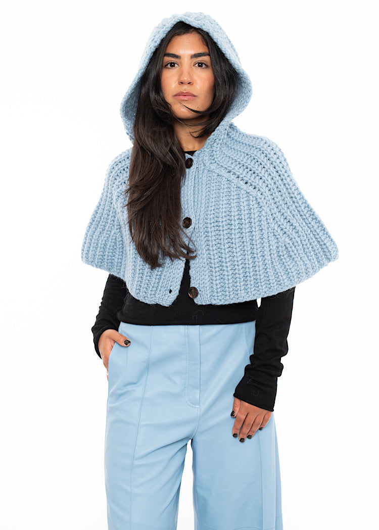 A model wearing an oversized knitted shawl in light blue. With the hood up showing the size and fit off the hood.