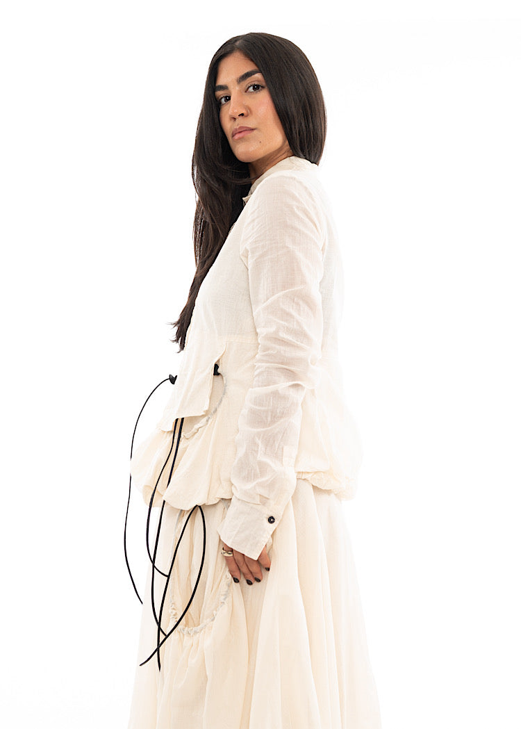 A Model standing to the side wearing a sculptural Jacket with a Skirt in the matching light beige colour. Focusing on the width and fit of the Jacket.