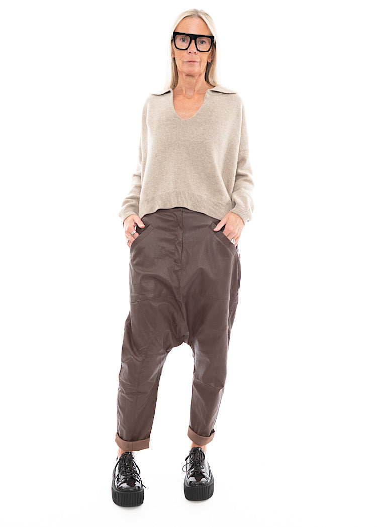 A full frame photo of A Model wearing a Structured Drop-Crotch Brown Leather Trouser, Disclosing the pockets.Styled With a patent Brown Shoe and a creme pullover.