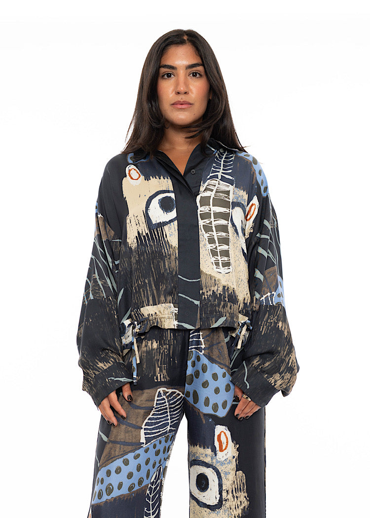 A model wearing an oversized silk shirt with multicoloured print throughout. A detailed hidden button effect to fasten, slightly tucked in to a matching silk wide leg trouser to add volume paired with black high heels.