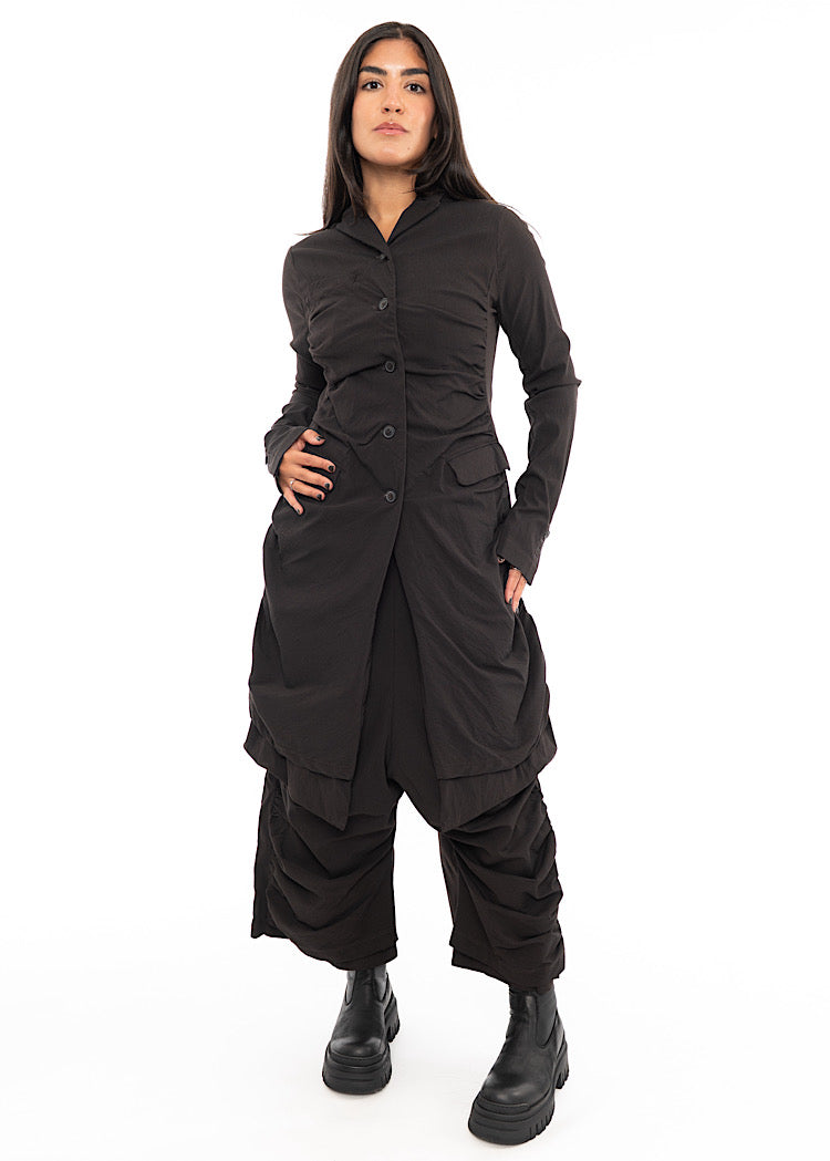 A full frame photo of A Model wearing a detailed techno coat and matching trousers, in a dark off-Brown tone. Paired with a chunky Boot.