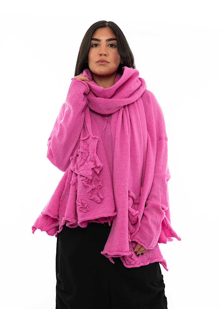 A Model wearing a Bright Pink Scarf with ruffled details surrounding the bottom of the piece. Paired with a matching pullover and Black Trouser.